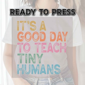 May include: A white t-shirt with the text "It's a good day to teach tiny humans" in colorful, distressed lettering. The text is in a rainbow color scheme.