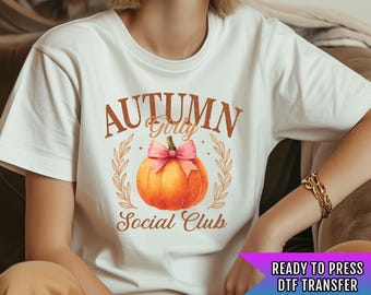 Pode incluir: Camiseta branca com um gráfico de abóbora marrom e laranja e o texto "Autumn Girly Social Club". A abóbora tem um laço rosa no topo. O texto está em uma fonte de estilo vintage.