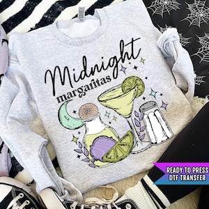 May include: A gray sweatshirt with the text "Midnight margaritas" in black lettering. The design includes a crescent moon, a bottle, a margarita glass, a lime wedge, and two salt shakers.