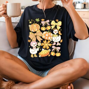 May include: Black t-shirt featuring a colorful fall-themed graphic. The design includes pumpkins, bows, flowers, and cartoon ducks. The pumpkins have orange and white patterns. The bows are in shades of yellow, pink, and orange. The ducks are wearing boots and hats.