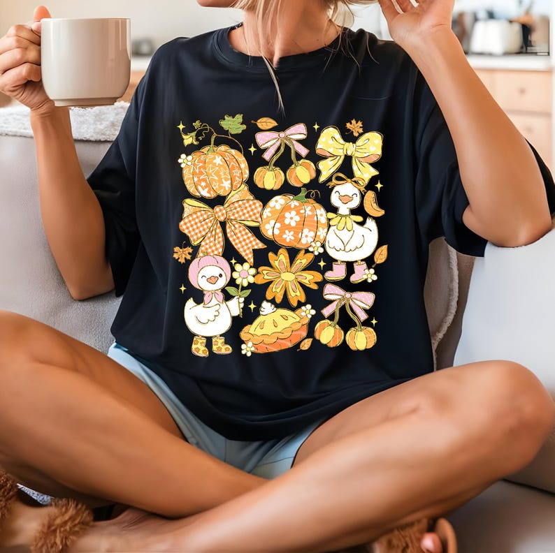 May include: Black t-shirt featuring a colorful fall-themed graphic. The design includes pumpkins, bows, flowers, and cartoon ducks. The pumpkins have orange and white patterns. The bows are in shades of yellow, pink, and orange. The ducks are wearing boots and hats.