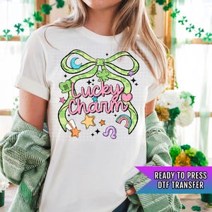 May include: White t-shirt featuring a green bow design with the words "Lucky Charm" in pink and green. The design includes stars, a rainbow, a horseshoe, and a shamrock. The text "READY TO PRESS DTF TRANSFER" is in the bottom right corner.
