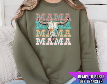May include: A green sweatshirt with a distressed, layered design that says "Mama" with a skull and crossbones in the center. The design is in shades of pink, yellow, and blue. The text "Ready to Press DTF Transfer" is at the bottom of the image.