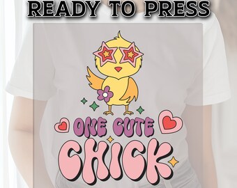 May include: A white t-shirt featuring a cartoon chick wearing star-shaped sunglasses. The chick holds a flower and is surrounded by hearts and stars. Text on the shirt reads "ONE CUTE CHICK" and "READY TO PRESS".