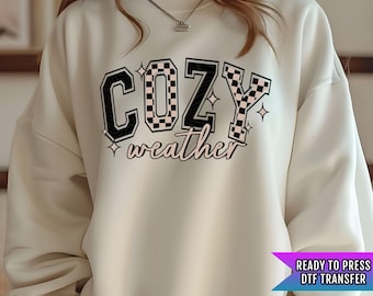 May include: A white sweatshirt with a black and white checkered graphic that says "Cozy weather" in pink lettering.