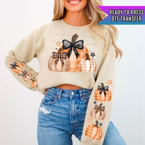 May include: A beige sweatshirt with a design of three pumpkins with black and white bows and stars. The pumpkins are orange and white with polka dots and plaid patterns. The text "READY TO PRESS DTF TRANSFER" is in the upper right corner of the image.