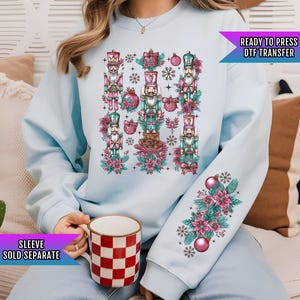 May include: A light blue sweatshirt with a Christmas-themed design featuring multiple nutcracker figures, snowflakes, and floral elements. The text "READY TO PRESS DTF TRANSFER" is visible in the top right corner of the image.