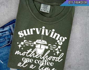 Könnte beinhalten: Ein grünes T-Shirt mit einer weißen Grafik, auf der "surviving motherhood one coffee at a time" steht. Die Grafik enthält auch eine Kaffeetasse mit einem Strohhalm und die Worte "always tired".