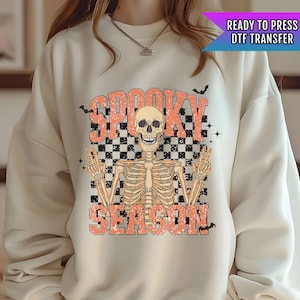 May include: A cream colored sweatshirt with a graphic print of a skeleton with the text "Spooky Season" in orange. The skeleton is wearing a checkered pattern behind it. The text "Ready to Press DTF Transfer" is in a blue and purple box in the top right corner.