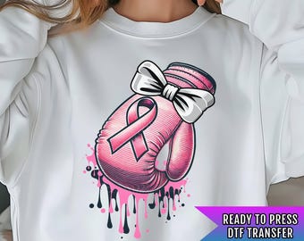 May include: A white sweatshirt with a pink boxing glove design. The glove has a pink ribbon tied around it with a white bow. The design is surrounded by pink and black paint splatters. The text "READY TO PRESS DTF TRANSFER" is at the bottom of the image.