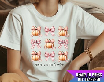 Pode incluir: Camiseta branca com um padrão repetido de abóboras em aquarela e laços rosa. As palavras "Pumpkin Patch Girly" são impressas abaixo das abóboras. O canto inferior direito tem o texto "READY TO PRESS DTF TRANSFER".