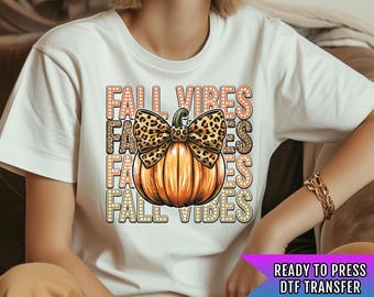 Pode incluir: Camiseta branca com um gráfico temático de outono. O design apresenta uma abóbora laranja com um laço de estampa de leopardo e as palavras "FALL VIBES" empilhadas. Transferência DTF pronta para prensar.