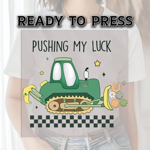 May include: A green and yellow cartoon bulldozer with the text "Pushing My Luck" and a checkered pattern at the bottom. The bulldozer is surrounded by stars, horseshoes, and four-leaf clovers.
