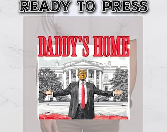 May include: White t-shirt with a graphic print of a man in a suit standing in front of the White House. The text "DADDY'S HOME" is printed above the image.