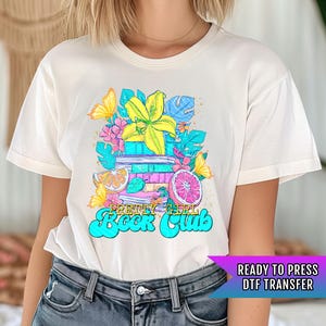 May include: White t-shirt with a colorful graphic design featuring a stack of books, flowers, and fruit. The text "Pretty Girl Book Club" is written in a retro font.
