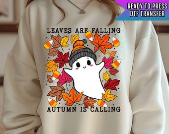 Pode incluir: Uma camisa de moletom branca com um design de fantasma fofo. O fantasma está cercado por folhas de outono coloridas e milho doce. O texto "Leaves are falling, autumn is calling" está impresso na camisa de moletom.