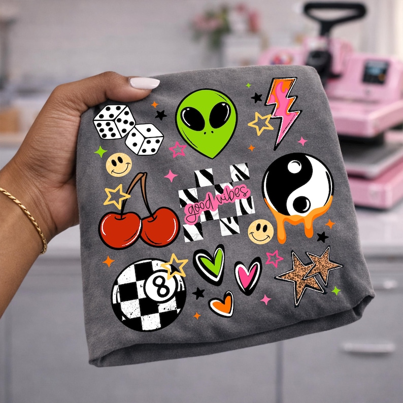 May include: A dark gray fabric featuring a colorful graphic print of dice, a green alien head, a pink lightning bolt, cherries, a yin-yang symbol, a checkered pattern with the text good vibes, smiley faces, hearts, stars, and an eight ball.