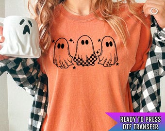 May include: Orange t-shirt featuring three cartoon ghosts with black outlines and star accents. One ghost has a checkered pattern. The text "READY TO PRESS DTF TRANSFER" is in a purple and blue banner.