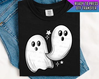 May include: Black t-shirt featuring a graphic of two white cartoon ghosts with black eyes and a star design. The shirt is folded on top of blue denim jeans. The text "READY TO PRESS DTF TRANSFER" is visible.
