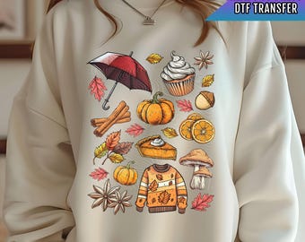 Pode incluir: Uma camisa de moletom bege com um design temático de outono colorido. O design inclui uma abóbora, uma torta, um cupcake, um guarda-chuva, paus de canela, folhas e um cogumelo. O design é perfeito para o outono.