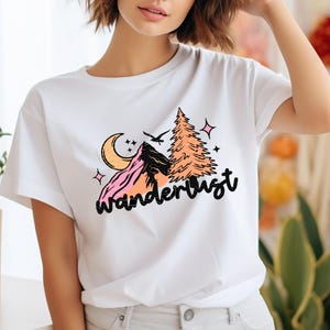 May include: White t-shirt with a graphic design of a mountain range, a pine tree, a crescent moon, and stars. The text "wanderlust" is written in a black cursive font.