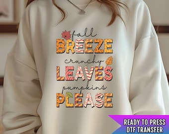 Pode incluir: Uma camisa de moletom branca com um design gráfico que diz "fall breeze crunchy leaves pumpkins please" em letras laranja, amarelo e marrom com um padrão de folhas de outono. O texto está pronto para ser estampado como uma transferência DTF.