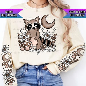 May include: A cream-colored sweatshirt with a raccoon, mushrooms, flowers, and a crescent moon graphic. The text "stay wild" is printed on the sweatshirt. The sleeves have a repeating pattern of mushrooms, flowers, and a crescent moon.