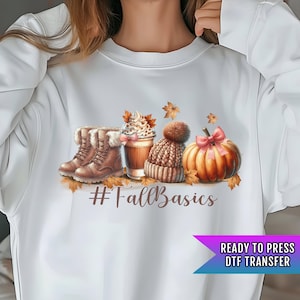 May include: White sweatshirt with a graphic design of a pumpkin, a cup of coffee, a knitted hat, and boots. The text "#FallBasics" is printed below the graphic.