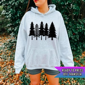 May include: A light gray hoodie featuring a black graphic of five stylized pine trees. The hoodie has a front pocket and a drawstring hood. The text "READY TO PRESS DTF TRANSFER" is displayed on a purple banner.
