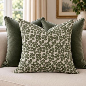 May include: A square throw pillow featuring a cream-colored fabric with a dense, repeating pattern of small, dark olive green floral shapes. This pillow is positioned in front of two solid, dark olive green velvet-textured throw pillows.