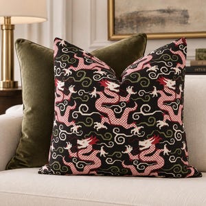 May include: A square throw pillow featuring a black background with a repeating pattern of red and white dragons interspersed with green and white swirling cloud motifs. Behind it sits a solid olive green velvet throw pillow.