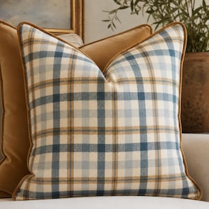 May include: A square throw pillow featuring a plaid pattern in shades of blue, tan, and cream. The pillow is finished with a solid tan velvet-like piping along the edges, providing a structured border to the soft, woven fabric design.