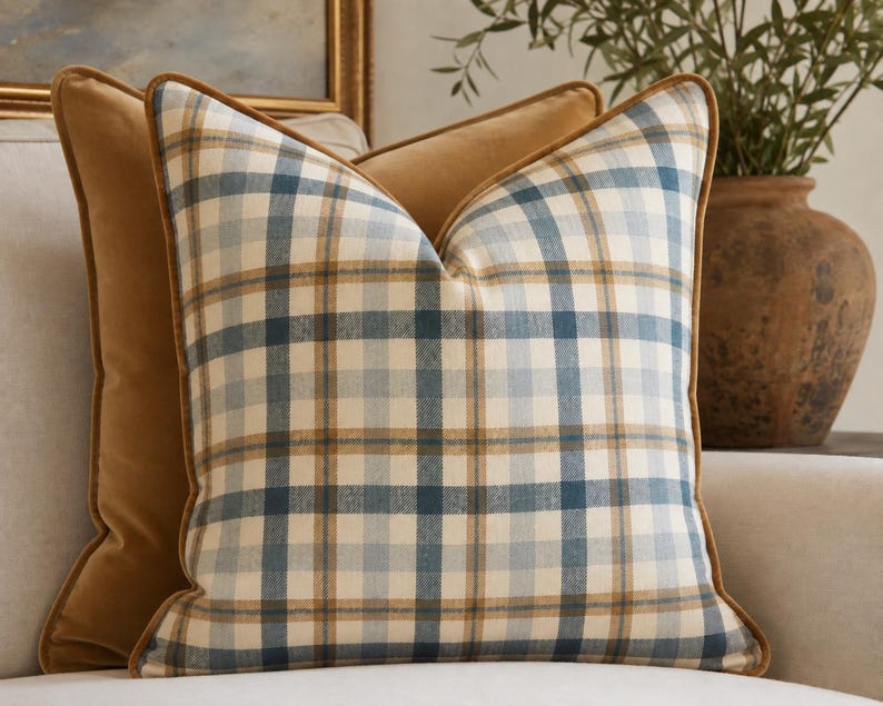 May include: A square throw pillow featuring a plaid pattern in shades of blue, tan, and cream. The pillow is finished with a solid tan velvet-like piping along the edges, providing a structured border to the soft, woven fabric design.