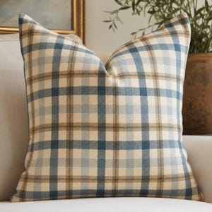 May include: A square throw pillow featuring a plaid pattern with intersecting lines in shades of cream, muted blue, and tan. The fabric has a woven texture, and the pillow is plumped to create a structured shape with defined corners and edges.