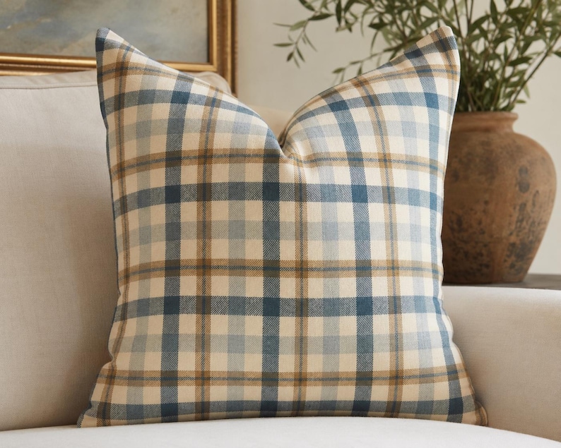May include: A square throw pillow featuring a plaid pattern with intersecting lines in shades of cream, muted blue, and tan. The fabric has a woven texture, and the pillow is plumped to create a structured shape with defined corners and edges.