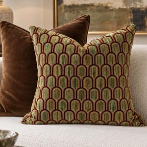 May include: A square throw pillow featuring a repeating arch pattern in shades of green, brown, and tan, positioned in front of a solid dark brown velvet throw pillow. Both pillows have a soft, plush texture and are square in shape.