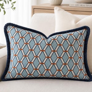 May include: Decorative lumbar pillow with a geometric pattern in shades of blue, white, and rust. The pillow has a dark blue fringe trim and is placed on a cream-colored sofa. A second cream-colored pillow is behind it.