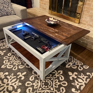 May include: A rectangular coffee table with a white wooden base and a dark brown stained wooden top that slides open to reveal a hidden storage compartment. The interior features a black foam-lined tray with LED lighting, holding two handguns.