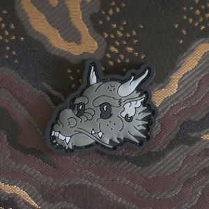 Puede incluir: Un parche bordado gris y negro de la cabeza de un dragón de dibujos animados con un contorno blanco. El parche está sobre un fondo de tela de camuflaje.