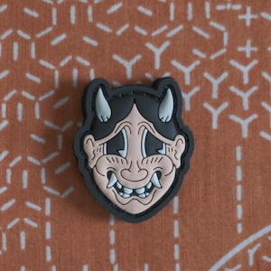 May include: A black and white rubber patch with a cartoon image of a demon with horns, a wide smile, and large eyes. The patch is on a brown and white patterned background.