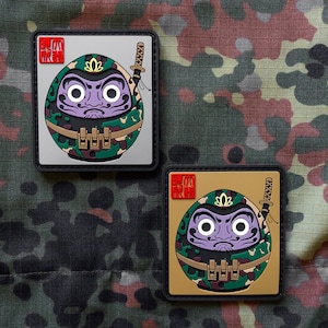 Peut inclure: Deux patchs carrés avec un personnage de dessin animé au centre. Le personnage est un daruma rond, vert et marron avec un visage violet et un masque de ninja noir. Le daruma tient une épée. Les deux patchs sont entourés d'une bordure noire. Le patch du haut a un carré rouge avec des caractères japonais dans le coin supérieur gauche. Le patch du bas a un fond doré et un carré rouge avec des caractères japonais dans le coin supérieur gauche.