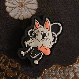 May include: A black and white cartoon dog-shaped rubber charm with a pink tongue and ears. The charm is on a dark background with a floral pattern.