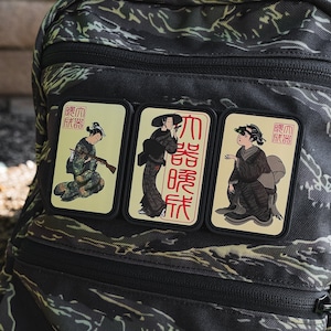 Puede incluir: Tres parches rectangulares con arte japonés en una mochila de camuflaje. Cada parche representa a una geisha con vestimenta tradicional, una sosteniendo un rifle y otra con texto japonés. Los parches tienen bordes negros.