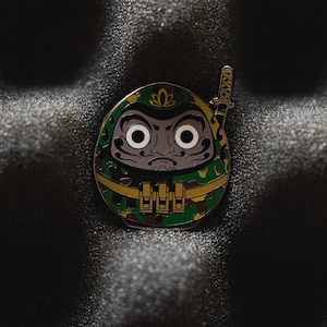 May include: A green and gold enamel pin of a cartoon character with a camouflage pattern and a sword. The character has a large head with a serious expression and large eyes.