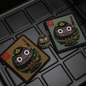 May include: Three items with a Daruma doll design in a camouflage pattern. Two square patches and a small keychain are visible. The patches feature Japanese text and a sword graphic. The items are on a black, textured surface.