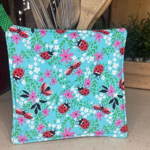 Può includere: Un sottopentola blu e verde con un motivo a coccinella e fiori. Il sottopentola ha un nastro verde e bianco.