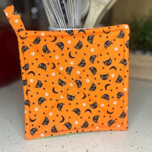 May include: Orange and black quilted hot pad with a cat and crescent moon pattern.