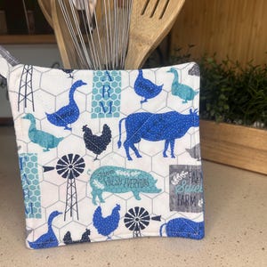 Puede incluir: Un agarrador de olla acolchado azul y blanco con un tema de animales de granja. El agarrador de olla presenta un fondo blanco con animales de granja azules y verdes, incluidos patos, pollos, un cerdo y una vaca. El texto "Farm Sweet Farm" está impreso en el agarrador de olla.