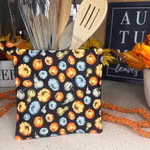 May include: A black pot holder with a fall-themed print of gourds and pumpkins in shades of orange, blue, and green. The pot holder has a loop handle.