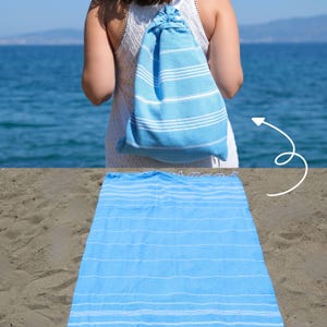 Pode incluir: Uma toalha de praia azul clara com listras brancas está estendida na areia. Uma mochila a condizer com cordão é usada por uma pessoa ao fundo. A toalha e a mochila são feitas de tecido leve.